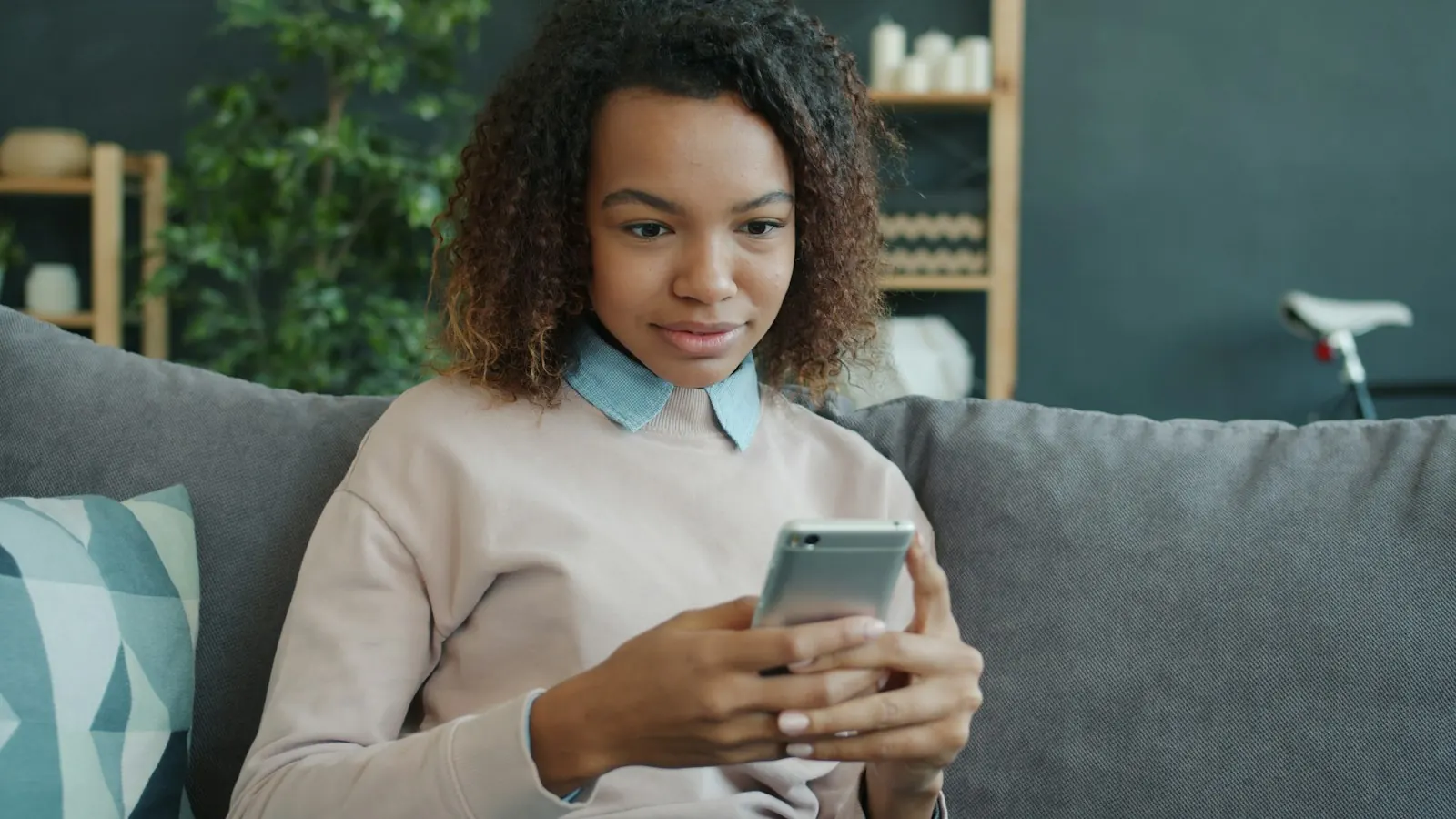 A young woman sits on a sofa at home, reading something on her phone